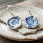 Gros plan de boucles d’oreilles artisanales en faïence recyclée bleue et blanche, posées sur des fragments d’assiette ancienne dans une lumière naturelle douce.