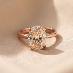 Bague élégante en or rose sertie d’un diamant champagne ovale, photographiée sur un fond beige avec une lumière naturelle douce.